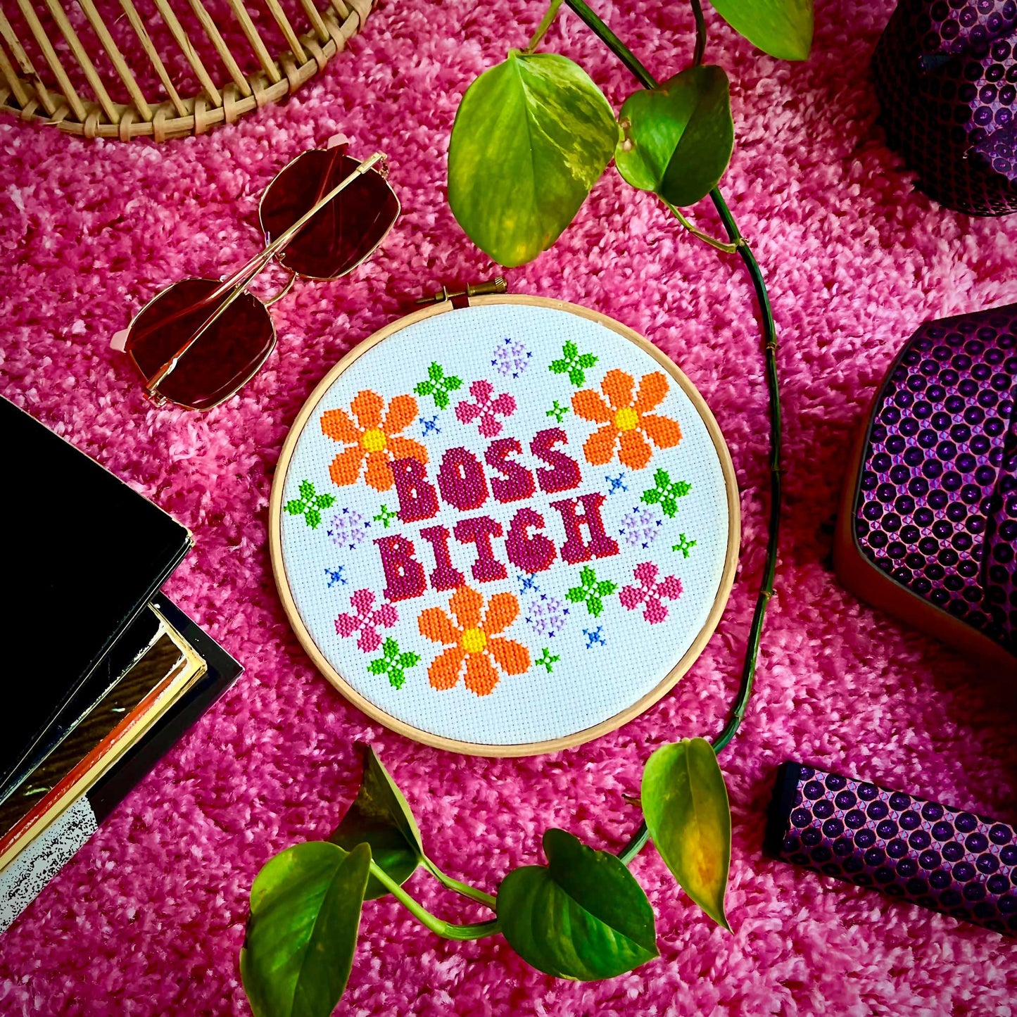 A 70's style cross stitch of the words 'boss bitch' decorated with flowers is laid on a fuzzy pink carpet surrounded by 70's style items
