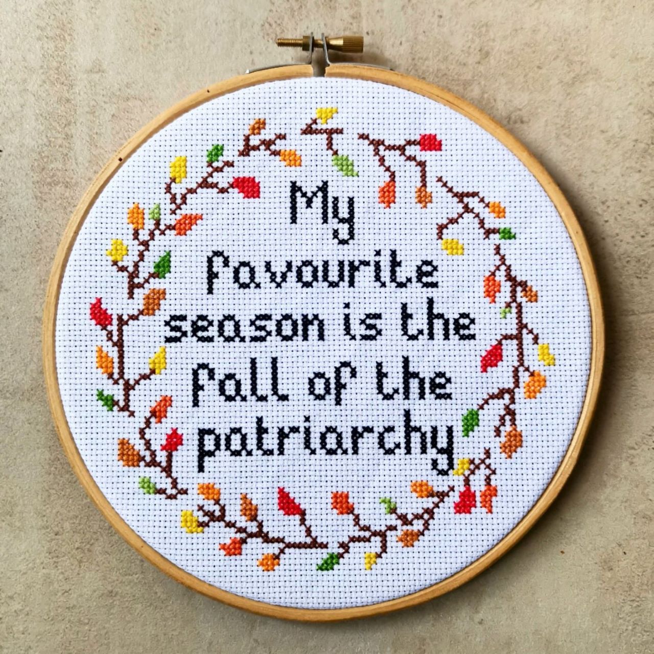 A close up of a cross stitch of the words 'My favourite season is the fall of the patriarchy' inside a wreath of autumn leaves, set inside a wooden embroidery hoop and on a stone background.