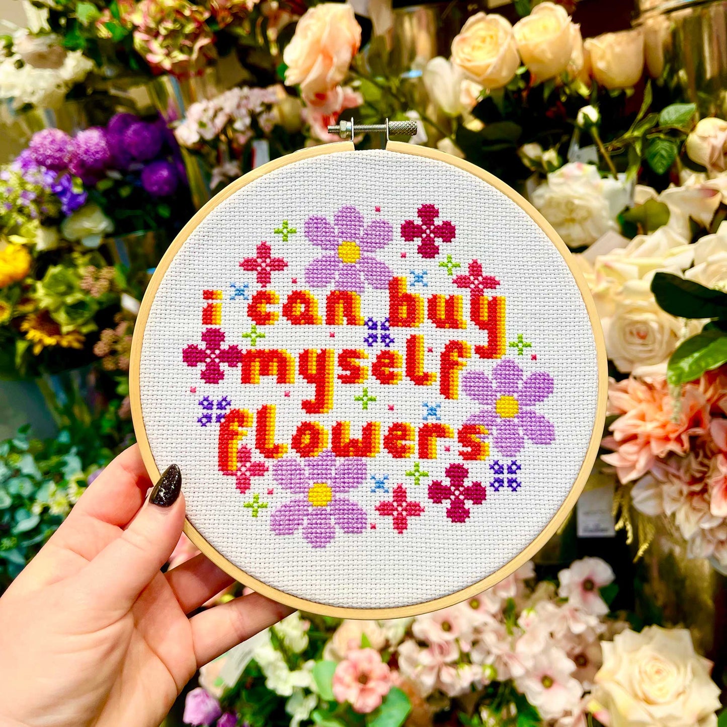 A cross stitch of the words I can buy myself flowers surrounded by stitched flowers is held up in front of a flower stall.
