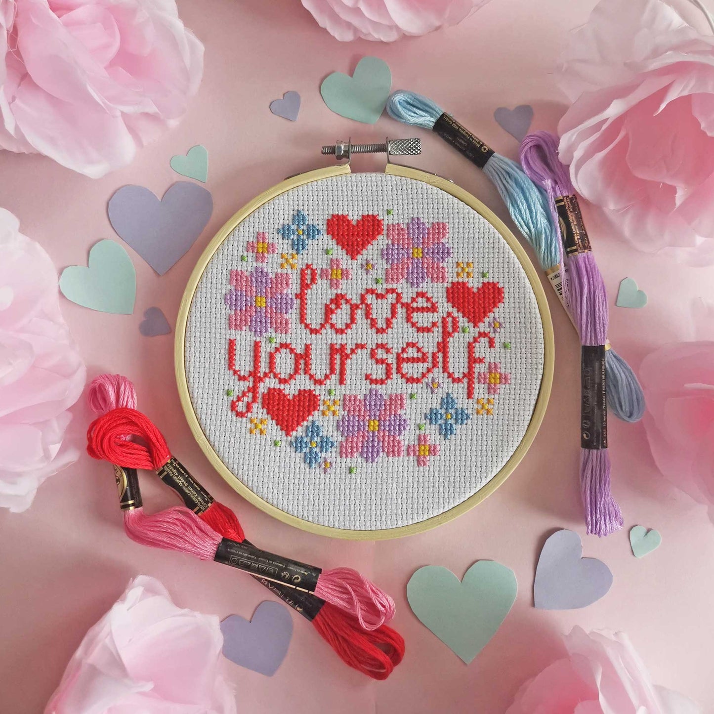 A cross stitch of the words love yourself and flowers and hearts is on a pink background surrounded by colourful paper hearts and fake pink roses.