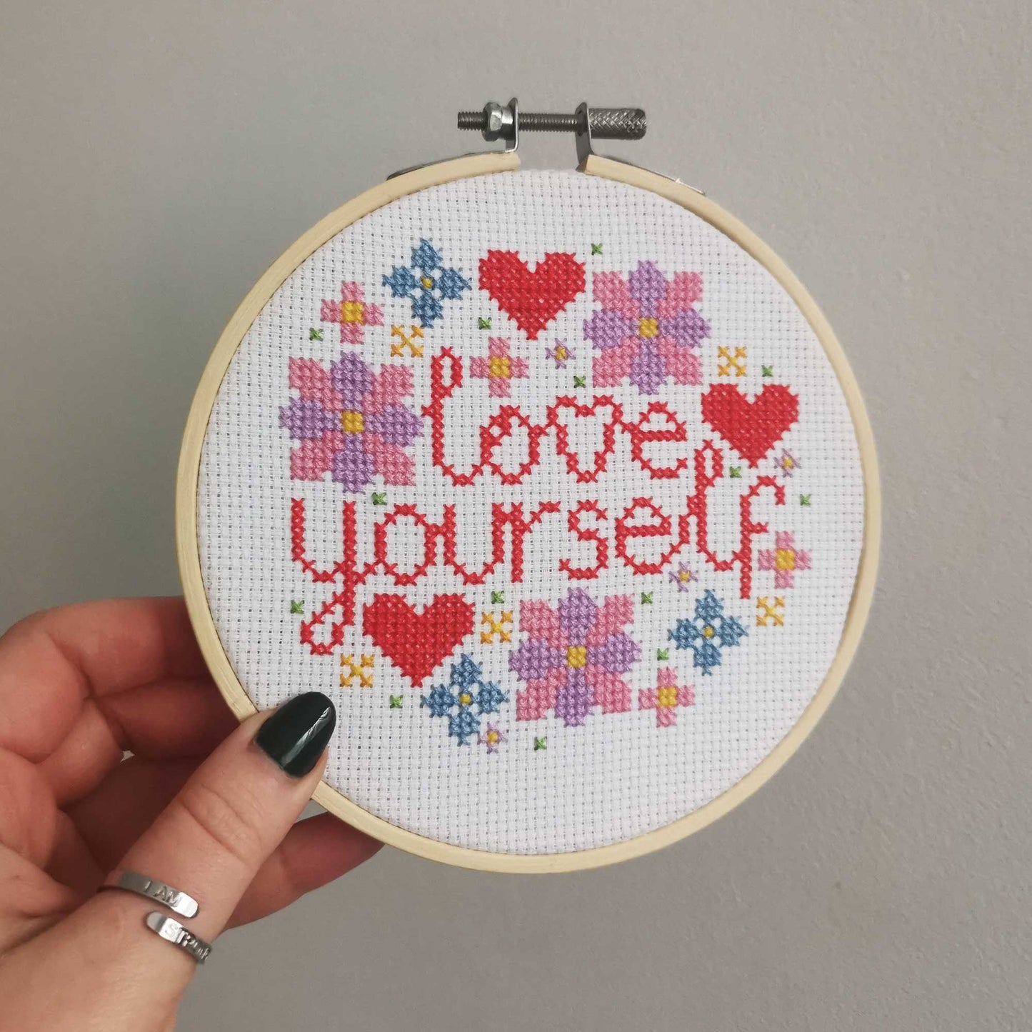 A cross stitch of the words 'love yourself' with flowers and hearts is held up to the camera.