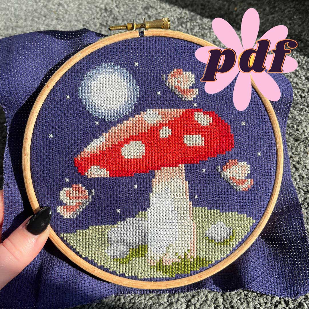 A cross stitch of a mushroom surrounded by moths, the moon and stars stitched on a dark blue fabric.