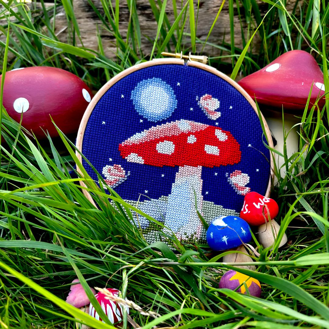A cross stitch of a mushroom with the moon, moths and stars nestled amongst grass.