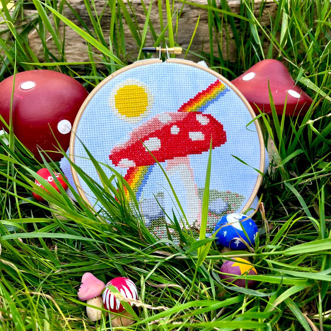 A cross stitch of a mushroom in front of a sun and rainbow is sat in grass surrounded by painted wooden mushrooms.