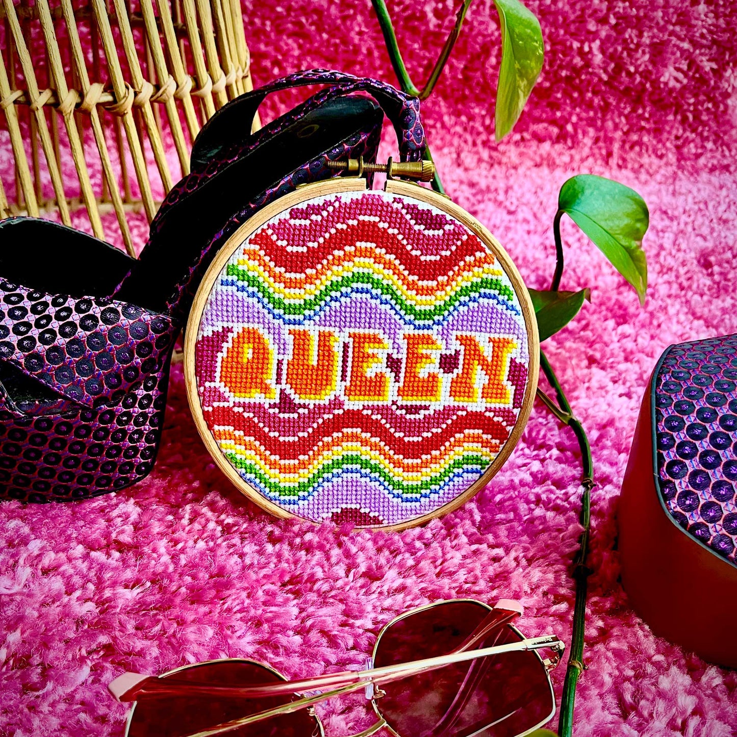 A wavy rainbow cross stitch leans against a platform heeled shoe with a bright pink carpet background.