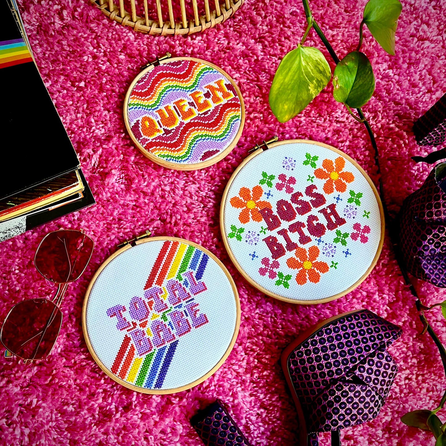 Three 1970's inspired finished cross stitches sit on a bright pink carpet, surrounded by records, sunglasses and shoes.