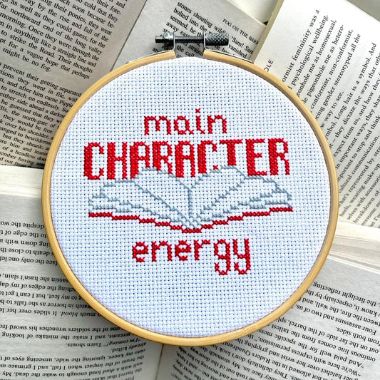 Cross-stitched hoop with 'main CHARACTER energy' text on an open book