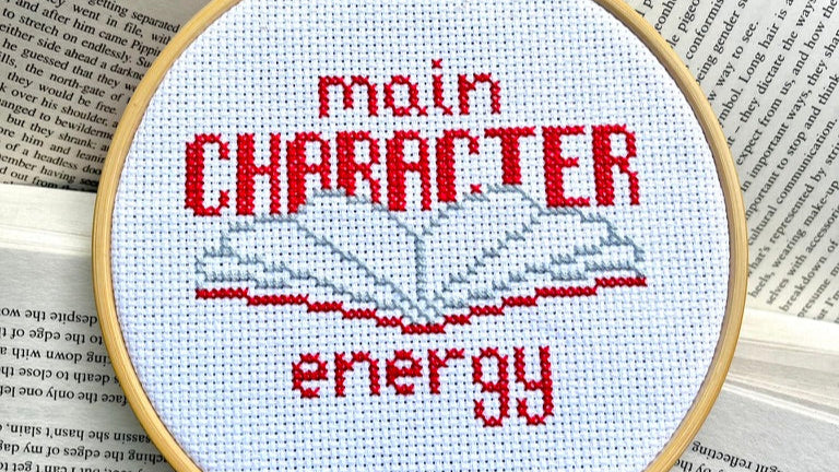 Cross-stitched hoop with 'main character energy' text on an open book
