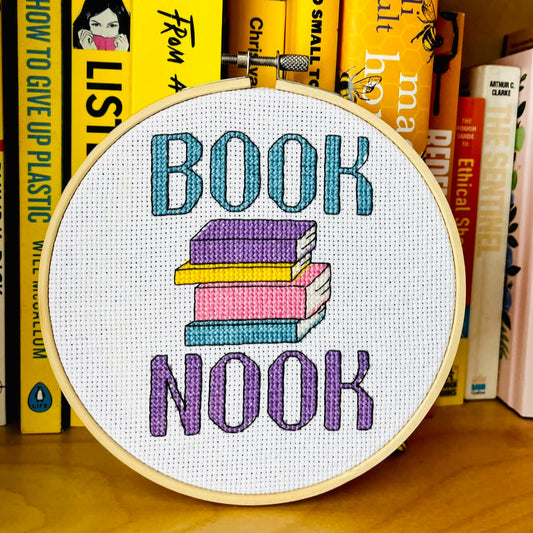Embroidery hoop with 'Book Nook' text and book illustration cross stitch against a bookshelf background