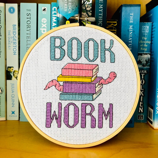 Embroidery hoop with 'Book Worm' cross stitch design on a bookshelf background