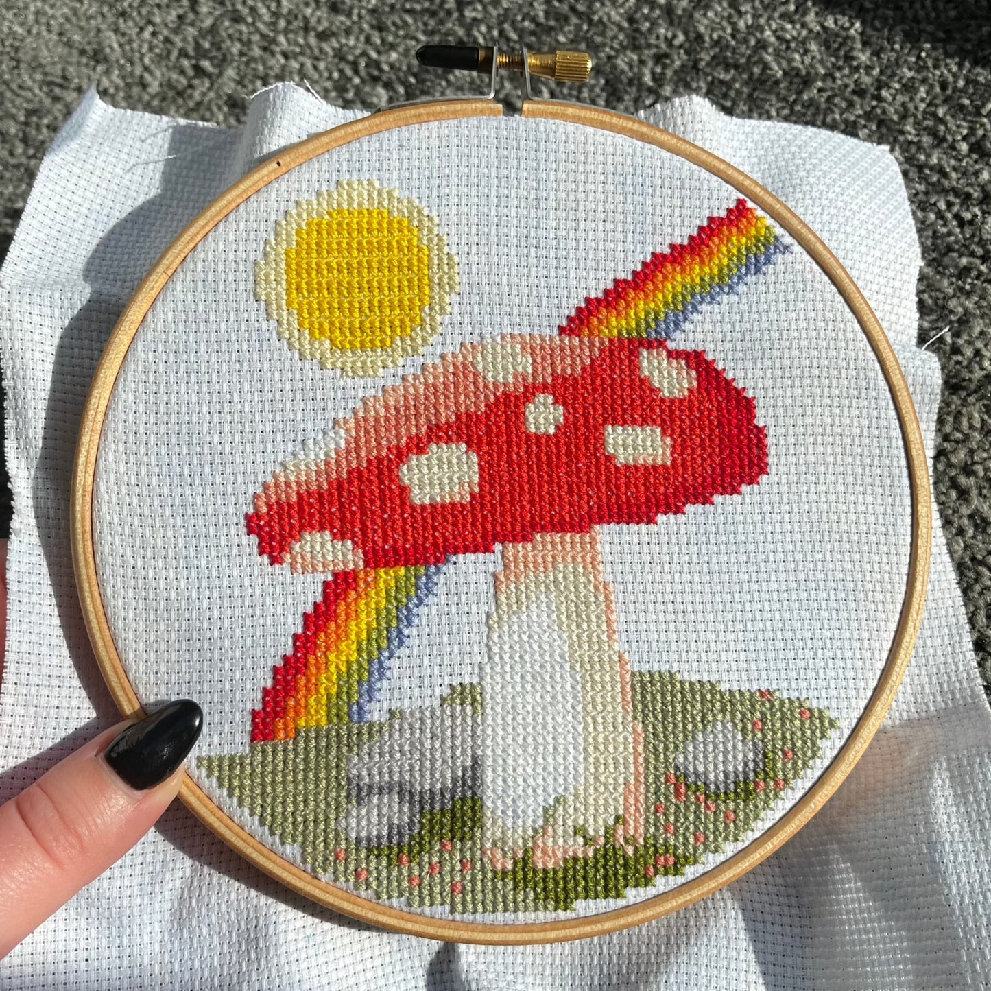 A cross stitch of a mushroom, sun and rainbow in a wooden hoop frame is held close to the camera
