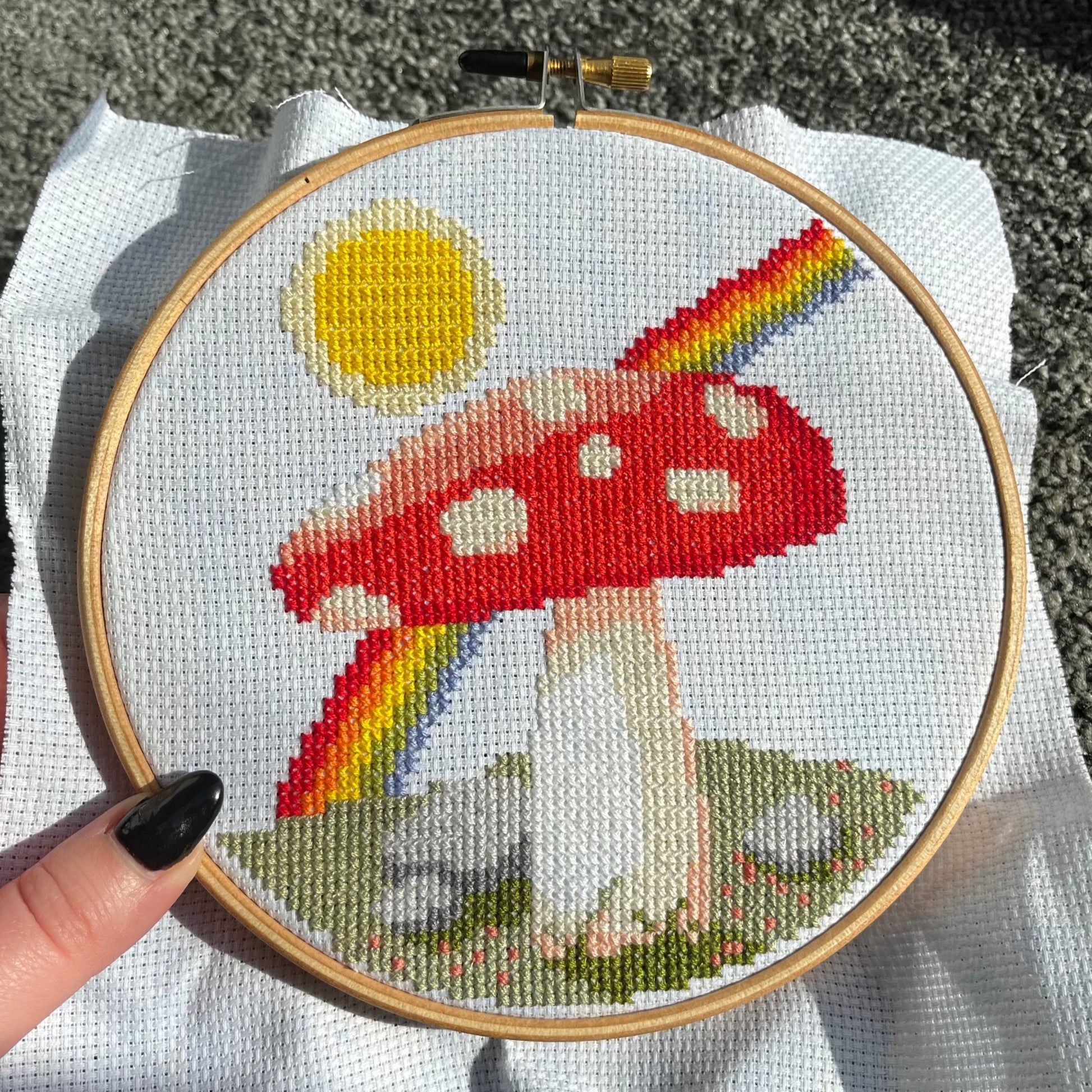 A cross stitch of a mushroom, sun and rainbow in a wooden hoop frame is held close to the camera
