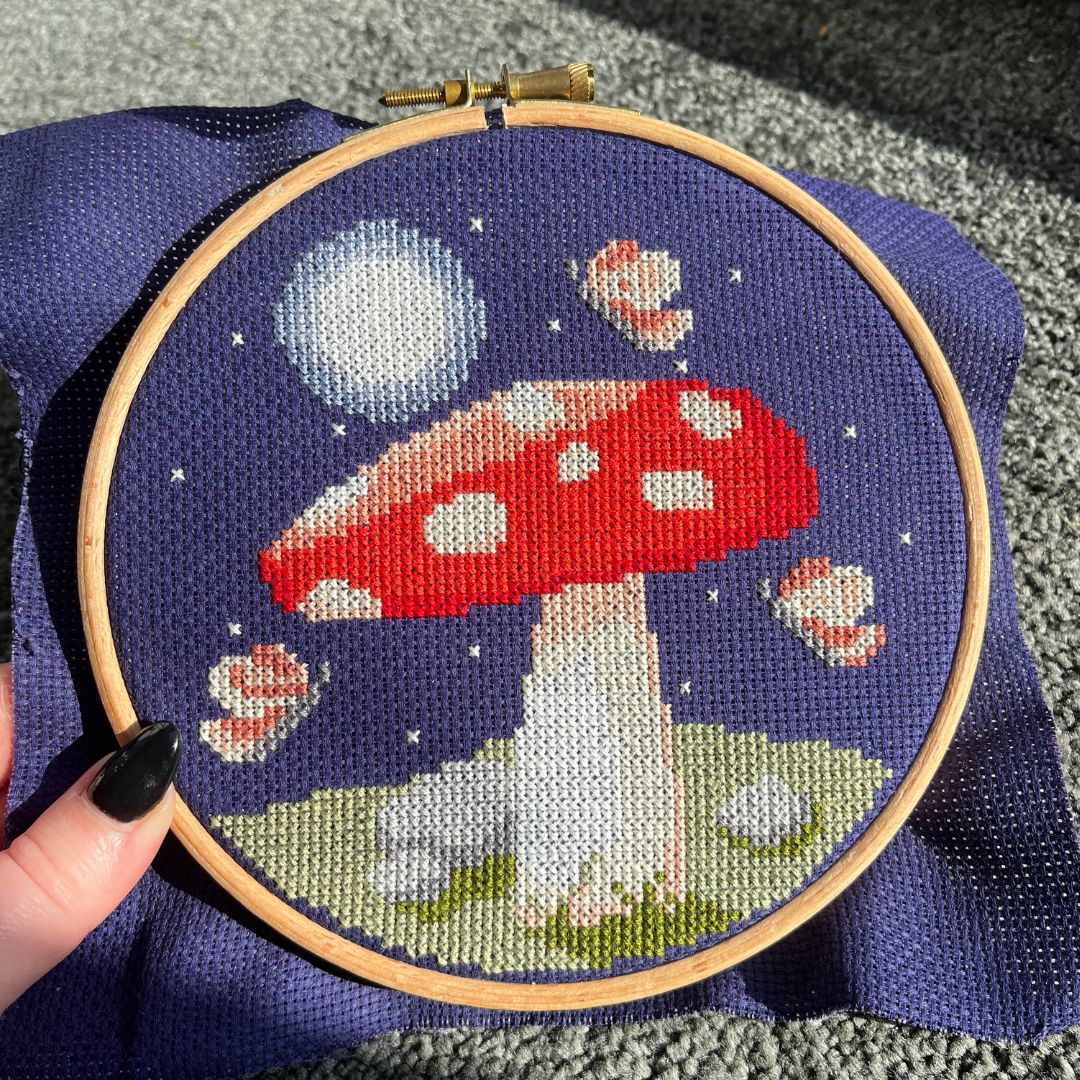 Close up of a cross stitch of a mushroom at night framed in a wooden embroidery hoop.